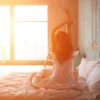 Woman stretching peacefully in a sunlit bedroom in morning light representing restful sleep and healthy circadian rhythms